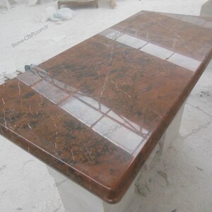 May include: A rectangular, polished countertop made of reddish-brown granite. The surface has dark veining and a glossy finish, with a lighter-colored inset section. The countertop is likely for a kitchen or bathroom.