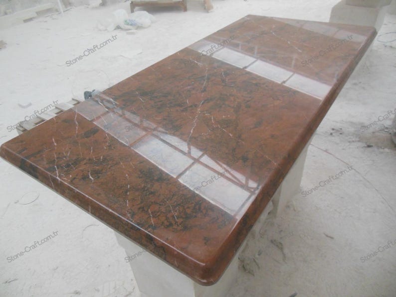 May include: A rectangular, polished countertop made of reddish-brown granite. The surface has dark veining and a glossy finish, with a lighter-colored inset section. The countertop is likely for a kitchen or bathroom.