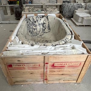 May include: An oval-shaped bathtub carved from white marble with prominent black veining, secured inside a wooden shipping crate. The crate features red stenciled text reading STONE-CRAFT.com.tr on the front panels and vertical support beam.