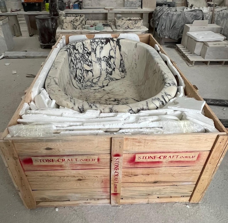 May include: An oval-shaped bathtub carved from white marble with prominent black veining, secured inside a wooden shipping crate. The crate features red stenciled text reading STONE-CRAFT.com.tr on the front panels and vertical support beam.