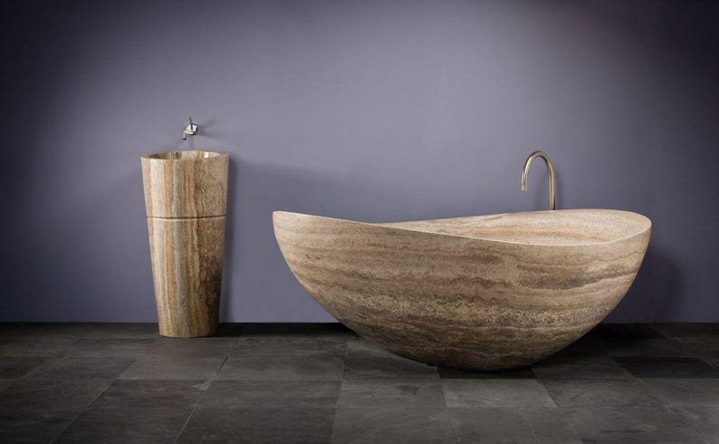 May include: A freestanding, oval-shaped bathtub and a matching cylindrical pedestal sink, both crafted from beige and brown veined natural stone. The bathtub features a curved, boat-like silhouette, and the sink includes a small metal faucet.