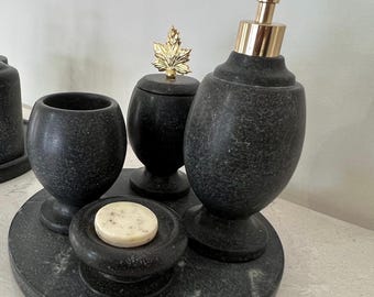 Könnte beinhalten: Ein Badezimmer-Set aus dunkelgrauem, gesprenkeltem Stein. Es besteht aus einem Seifenspender mit goldener Pumpe, einem Döschen mit goldenem Blatt-Knauf, einem Zahnputzbecher, einer Seifenschale mit weißem Seifenstück und einem runden Tablett.
