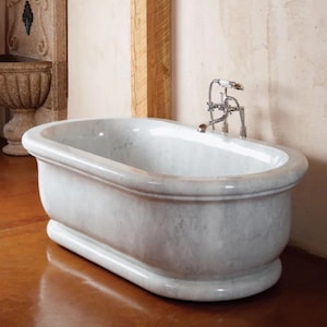 May include: A freestanding oval bathtub crafted from white marble with grey veining. The tub features a thick, rounded rim and a solid base. A polished chrome faucet assembly with two cross-style handles and a handheld shower attachment is mounted.