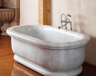 May include: A freestanding oval bathtub crafted from white marble with grey veining. The tub features a thick, rounded rim and a solid base. A polished chrome faucet assembly with two cross-style handles and a handheld shower attachment is mounted.
