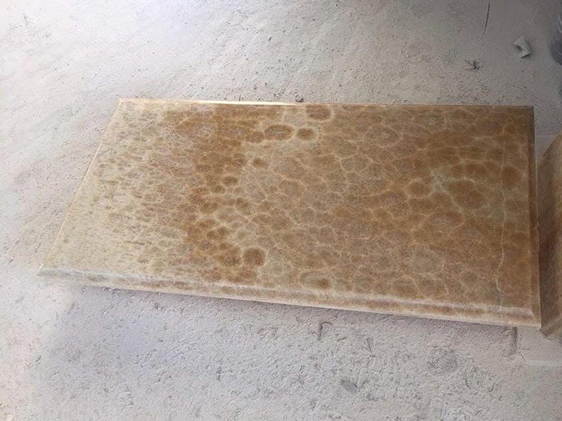 May include: A rectangular, light brown stone slab with a textured surface. The stone has a pattern of darker brown, irregular shapes across its surface. The edges of the slab are smooth and finished. The stone is likely for construction or design.