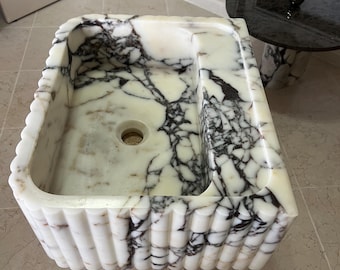 May include: A rectangular marble sink featuring a white base with prominent dark purple and black veining. The exterior walls of the basin have a vertical fluted texture, and the interior floor includes a centered circular drain opening.