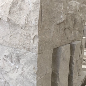 Op de afbeelding: Close-up van gestapelde, ruw bewerkte stenen blokken. De blokken zijn lichtgrijs van kleur met een textuur en zichtbare scheuren. De afbeelding toont de hoek van een groot blok en verschillende kleinere blokken.