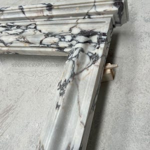 Puede incluir: Una repisa de chimenea de m&aacute;rmol blanco con vetas prominentes de color p&uacute;rpura oscuro y negro en toda la piedra. La estructura tiene un dise&ntilde;o de borde escalonado y moldeado, e incluye el texto STONE-CRAFT.com.tr impreso en la superficie frontal.