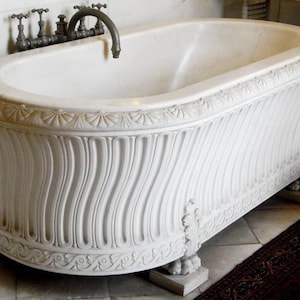 May include: A white marble bathtub featuring a carved fluted exterior, a decorative border with shell motifs along the top rim, and a base with ornate leaf carvings. The tub is equipped with a dark metal faucet assembly mounted on the wall.