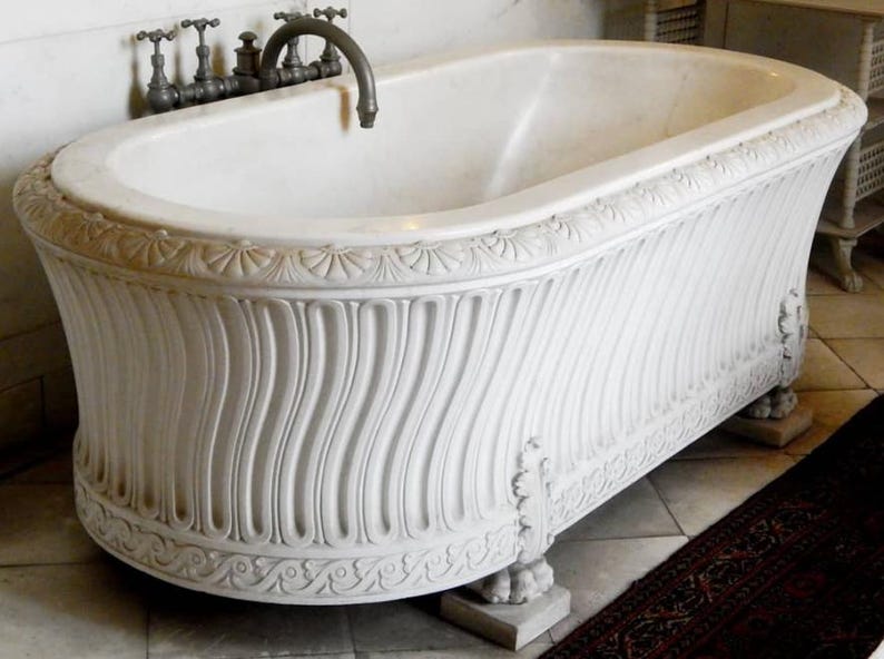 May include: A white marble bathtub featuring a carved fluted exterior, a decorative border with shell motifs along the top rim, and a base with ornate leaf carvings. The tub is equipped with a dark metal faucet assembly mounted on the wall.