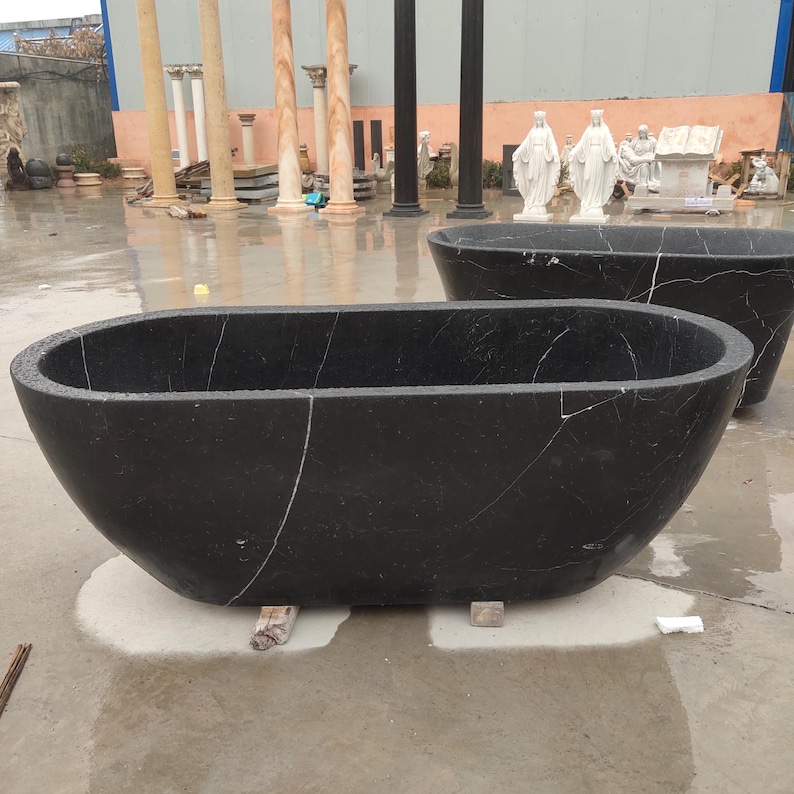 May include: A freestanding oval bathtub crafted from solid black marble with distinct white veining. The tub features a smooth, deep basin and a polished exterior surface, showcasing the natural stone texture and patterns throughout the material.