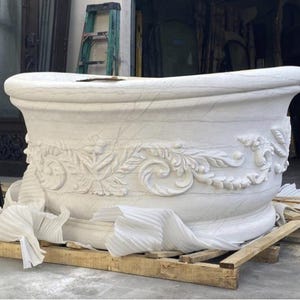 May include: A white stone bathtub featuring intricate carved floral and scroll relief patterns along the exterior side. The tub has a thick, rounded rim and a smooth, curved basin shape, resting on a wooden pallet with white protective foam.