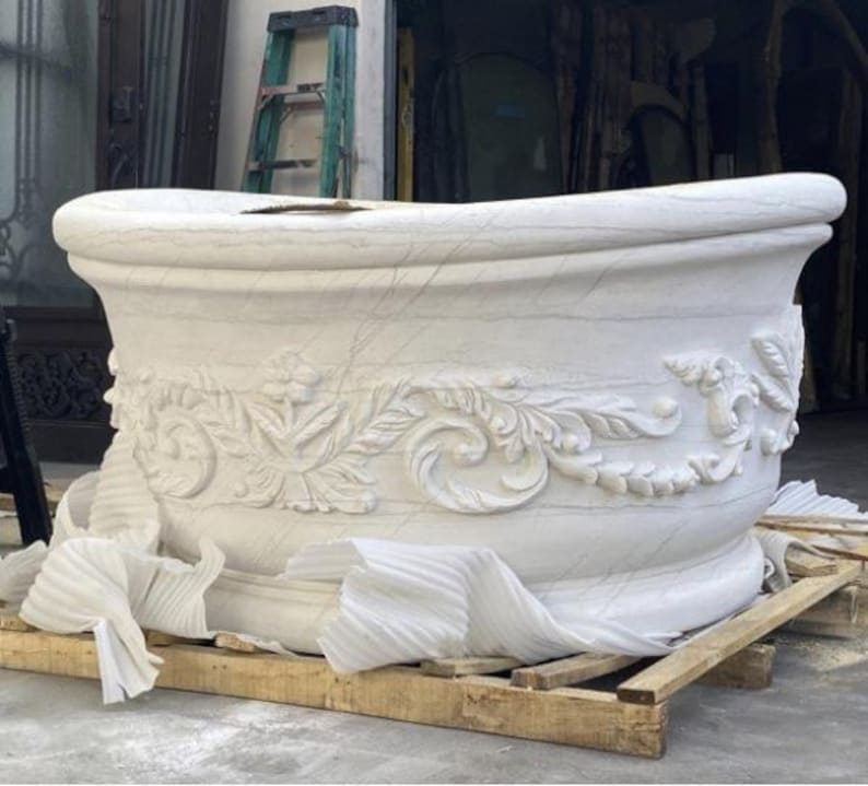 May include: A white stone bathtub featuring intricate carved floral and scroll relief patterns along the exterior side. The tub has a thick, rounded rim and a smooth, curved basin shape, resting on a wooden pallet with white protective foam.