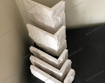Könnte beinhalten: Ein Stapel hellfarbiger, V-förmiger Steinstücke. Die Stücke haben eine raue, strukturierte Oberfläche und sind in einer versetzten Formation angeordnet. Der Stein scheint eine neutrale Farbe zu haben, möglicherweise cremeweiß oder hellbeige. Die Teile dienen wahrscheinlich Bau- oder Dekorationszwecken.
