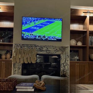 以下が含まれることがあります： 壁掛けテレビにはフットボールの試合が映し出され、その下にはベージュのニットストッキングが並ぶ暖炉の棚があります。暖炉はマーブルストーンで縁取られ、両サイドには様々な装飾品が置かれた木製の作り付け棚が設置されています。