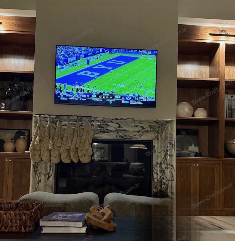 以下が含まれることがあります： 壁掛けテレビにはフットボールの試合が映し出され、その下にはベージュのニットストッキングが並ぶ暖炉の棚があります。暖炉はマーブルストーンで縁取られ、両サイドには様々な装飾品が置かれた木製の作り付け棚が設置されています。
