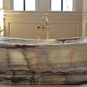 May include: A freestanding bathtub carved from natural banded onyx stone featuring horizontal layers of cream, green, and brown mineral deposits. The tub is equipped with a polished gold-tone metal faucet assembly with a handheld shower attachment.