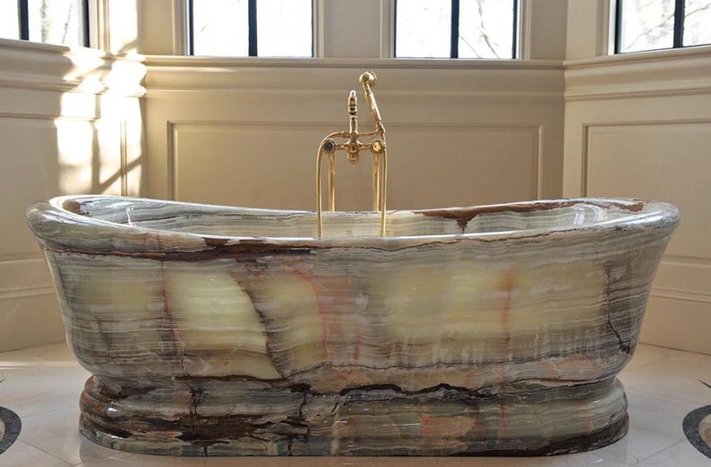 May include: A freestanding bathtub carved from natural banded onyx stone featuring horizontal layers of cream, green, and brown mineral deposits. The tub is equipped with a polished gold-tone metal faucet assembly with a handheld shower attachment.