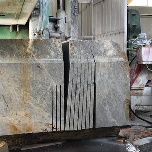 May include: A large, gray stone block with multiple parallel cuts, likely for construction or design. The stone has a rough, textured surface with some brown and yellow discoloration. Industrial machinery is visible in the background.