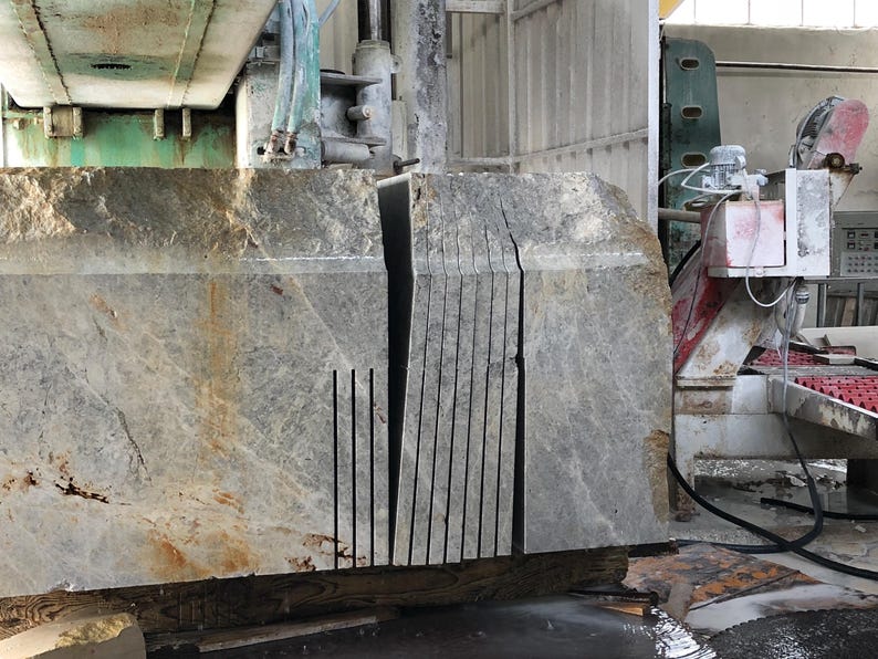 May include: A large, gray stone block with multiple parallel cuts, likely for construction or design. The stone has a rough, textured surface with some brown and yellow discoloration. Industrial machinery is visible in the background.