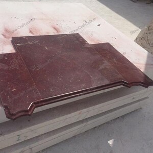 May include: A rectangular, burgundy-colored marble slab with a decorative edge. The stone has a polished surface and is set on a stack of lighter-colored stone blocks. The image is taken outdoors in natural light.