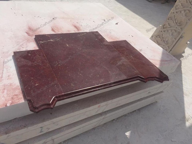 May include: A rectangular, burgundy-colored marble slab with a decorative edge. The stone has a polished surface and is set on a stack of lighter-colored stone blocks. The image is taken outdoors in natural light.