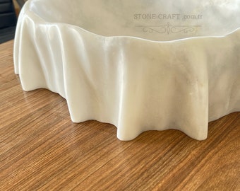 Könnte beinhalten: Ein weißes Waschbecken aus Marmor mit einem gewellten, stoffartigen Rand. Die Innenfläche zeigt den Text STONE-CRAFT.com.tr mit einer dekorativen Verzierung. Der Stein hat eine glatte, polierte Oberfläche mit dezenten grauen Maserungen.