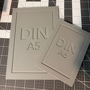 May include: Two rectangular gray plastic templates with beveled edges. The larger template features the embossed text DIN A5, while the smaller overlapping template features the embossed text DIN A6. Both items have a smooth, matte finish.
