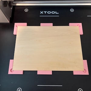 May include: A rectangular light-colored wooden board secured on a black xTool laser machine bed. The board is held in place by six pink plastic positioning jigs, with four L-shaped corner pieces and two rectangular side pieces, against a black surface.