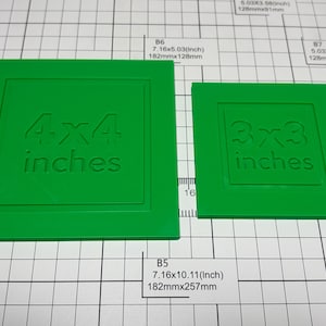 May include: Two green square plastic templates with embossed text. The larger template features the text 4x4 inches, and the smaller template features the text 3x3 inches. Both items are positioned on a white grid surface with measurement markings.