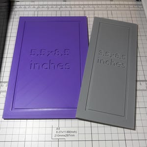 May include: A purple rectangular template with the text 5.5x8.5 inches and a grey rectangular template with the text 3.5x8.5 inches. Both templates feature a raised border design and are positioned on a white grid surface with measurement markings.