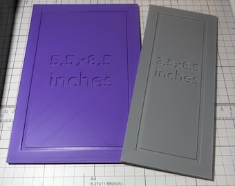 May include: A purple rectangular template with the text 5.5x8.5 inches and a grey rectangular template with the text 3.5x8.5 inches. Both templates feature a raised border design and are positioned on a white grid surface with measurement markings.