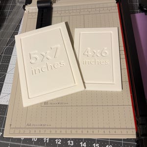 May include: Two off-white picture frames, one labeled "5x7 inches" and the other "4x6 inches", rest on a paper cutter. The frames have raised text. The paper cutter has a beige cutting surface with black and red accents.