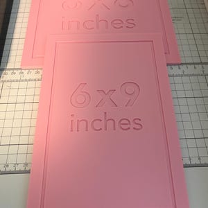 May include: Two rectangular pink silicone molds with raised borders. The top mold features the embossed text 8x8 inches, while the bottom mold features the embossed text 6x9 inches. Both molds are positioned on a white cutting mat with grid lines.