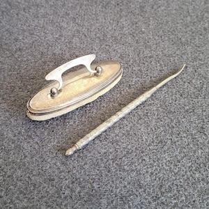 Op de afbeelding: Een zilveren nagelvijl met een klein, ovaal etui. Het etui heeft een zilveren handvat en een witte binnenkant.