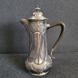 Op de afbeelding: Een zilveren koffiekan met een versierd ontwerp en een donkerbruine houten handgreep.