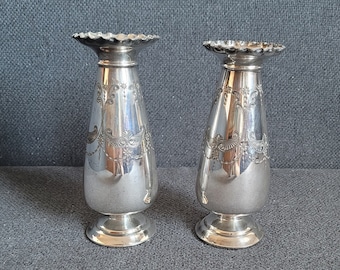May include: Two silver-colored, ornate vases with a flared, scalloped top and a bulbous body. The vases have a detailed floral design and sit on a round base. The background is a dark gray fabric.