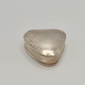 May include: A small, silver-coloured, heart-shaped box with a hinged lid. The box has a slightly rounded shape and is decorated with etched patterns along the edges and on the lid. The box is likely made of metal and has a polished finish.