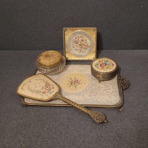 Könnte beinhalten: Ein vergoldetes Schminktablett mit floralem Design. Das Tablett enthält einen kleinen Spiegel, eine Uhr und zwei kleine Schachteln mit Blumenmustern.