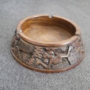 Op de afbeelding: Een ronde houten asbak met een gesneden reliëf ontwerp. De bruine asbak heeft een gedetailleerde gravure van dieren en bladeren aan de zijkanten. Het heeft een cirkelvormige inkeping voor het vasthouden van sigaretten.
