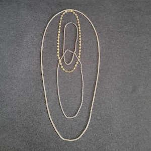 Może przedstawiać: Trzy złote i srebrne łańcuszki o różnych długościach, ułożone na szarej powierzchni tkaniny.