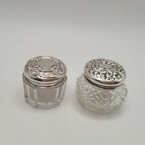 Puede incluir: Dos pequeños frascos de tocador con tapas de metal plateado con adornos en relieve. Un frasco tiene una base de vidrio transparente facetado con un patrón geométrico, mientras que el otro tiene una base de vidrio liso de forma hexagonal.