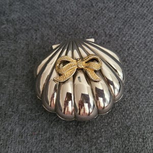 Könnte beinhalten: Eine silberfarbene Schmuckschatulle in Muschelform mit einer goldfarbenen Schleife. Die Schatulle hat eine polierte, reflektierende Oberfläche und ein detailliertes, geripptes Design. Die Schleife ist strukturiert und fügt ein dekoratives Element hinzu.