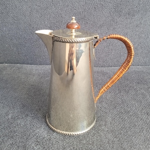 Puede incluir: Una cafetera de metal plateado con un cuerpo cilíndrico alto y un pico cónico. La tapa presenta un pequeño pomo redondo de color marrón. El asa es curva y está envuelta en un material marrón texturizado, con un detalle de cuerda en la base.