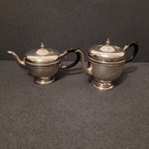 Puede incluir: Un juego de té de plata con una tetera y una jarra de crema. Ambas piezas tienen un cuerpo redondo con una tapa y un asa negra.