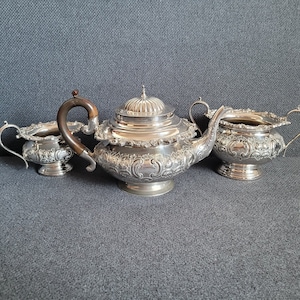 Könnte beinhalten: Ein silbernes Teeservice mit Teekanne, Milchkännchen und Zuckerdose. Die Teekanne hat einen Holzgriff und aufwendige Blumendekore. Auch das Milchkännchen und die Zuckerdose sind mit Blumenmustern verziert.