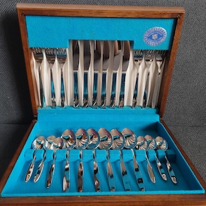 May include: A wooden box with a blue velvet lining contains a set of silver cutlery. The cutlery includes spoons, forks, and knives. The box has a silver label with the text "Stainless Steel"