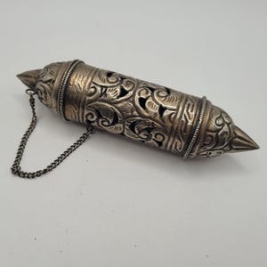 May include: Ornate silver-colored cylindrical container with intricate floral and scrollwork patterns. The container has pointed ends and a small chain attached. The design suggests it may be a decorative item or a holder for small items.