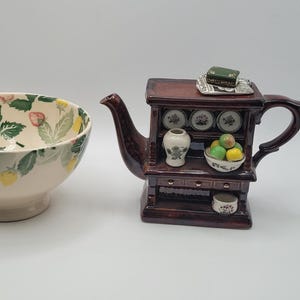 May include: A ceramic bowl and a teapot. The bowl has a cream-colored base with a strawberry and leaf pattern. The teapot is brown and shaped like a cabinet, with decorative plates, a vase, and a bowl of fruit.