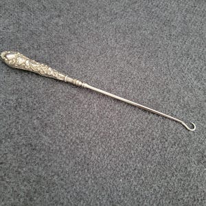 Può includere: Un uncinetto in metallo argentato con un manico ornato a motivi floreali. L'uncinetto si assottiglia fino a un fusto sottile e termina con un gancio curvo. Strumento per lavori di cucito e tessili.